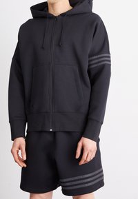 Svart zip-up hoodie med framfickor, dragsko för huvan och horisontella randiga detaljer. Kombinerad med svarta shorts med randiga detaljer.