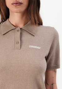 Beige strikket piké med kort erme, ribbet krage og to knapper. Har liten hvit brodert tekst "2ND DAY" på venstre side.