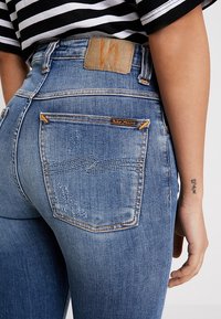 Džíny z modrého denimu s vysokým pasem a mírným vyblednutím. Mají oranžové prošívání, dvě zadní kapsy a koženou nášivku s logem na pase.