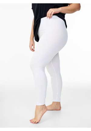 Vrouw draagt witte leggings tot op de grond en een zwart topje, staat op één voet op blote voeten tegen een effen witte achtergrond.