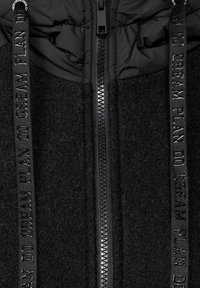 Chaqueta negra con cremallera que presenta un tejido texturizado, capucha suave e impermeabilizada y cordones decorativos con texto impreso.