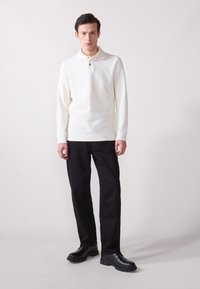 Camisa polo blanca de manga larga con un acabado texturizado, que presenta un cuello y un solo botón, combinada con pantalones negros de pierna recta y zapatos negros.
