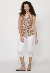Blusa senza maniche floreale con scollatura a V in bianco, rosso e verde; abbinata a pantaloni cropped bianchi con pizzo e decorazioni. Sandali neri.