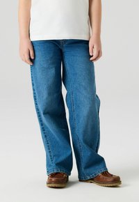 Barn iført vide blå jeans og brune skinnsko, stående mot en ensfarget lys bakgrunn.