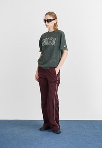 T-shirt oversized verde marmorizado com o logótipo "NIKE", combinado com calças burgundy com riscas brancas laterais e sapatos pretos slip-on.