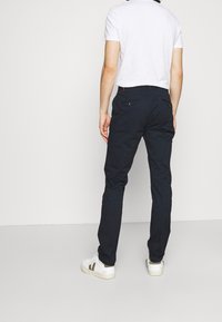 Pantaloni blu navy con un design a gamba dritta, dotati di due tasche posteriori con bottoni e una cintura elegante, abbinati a una t-shirt bianca e sneakers.