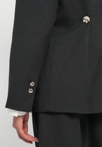 Person iført en mørkegrå dress med lysebrune knapper på ermet og jakken, hvit skjortemansjett synlig, hånd avslappet ved siden.