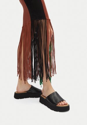 Czarne skórzane sandały na platformie z grubą podeszwą. Brązowe i czarne spodnie z ozdobnymi frędzlami, z zielonymi akcentami.