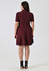Eine Frau steht mit dem Rücken zur Kamera und trägt ein knielanges burgunderrotes Kleid mit kurzen Ärmeln und schwarzen, hochhackigen Stiefeletten.