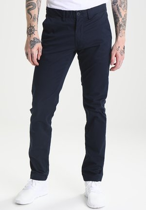 Homme portant un pantalon slim bleu marine foncé, des baskets blanches et une chemise blanche, debout avec les bras tatoués visibles.
