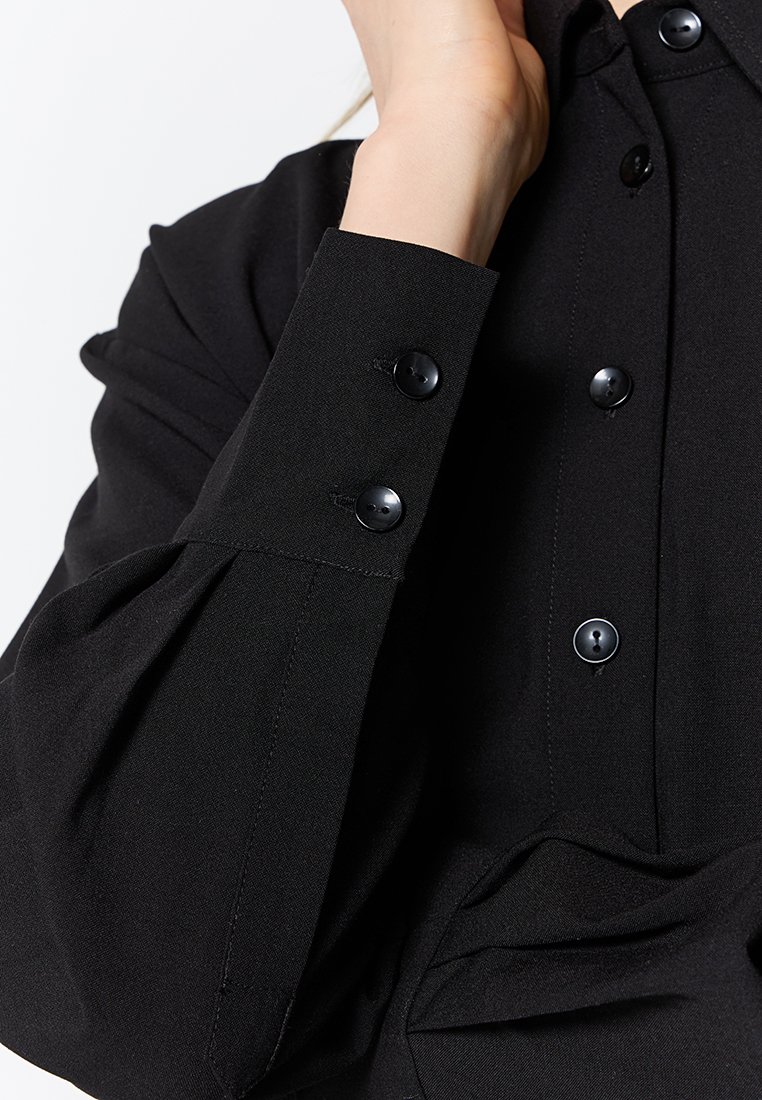 Schwarze Bluse mit geknöpften Manschetten, aus einem glatten, strukturierten Stoff. Das Design umfasst Falten und klassische schwarze Knöpfe als Detail.