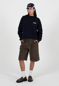 Femme portant un sweat-shirt bleu marine, un short marron arrivant aux genoux, des chaussettes blanches, des mocassins marron et une casquette bleu marine et bleu clair, debout les mains dans les poches.