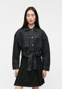 Chaqueta de denim negro con un diseño corto, botones y dos bolsillos frontales; presenta una cintura atada y costuras amarillas en contraste.