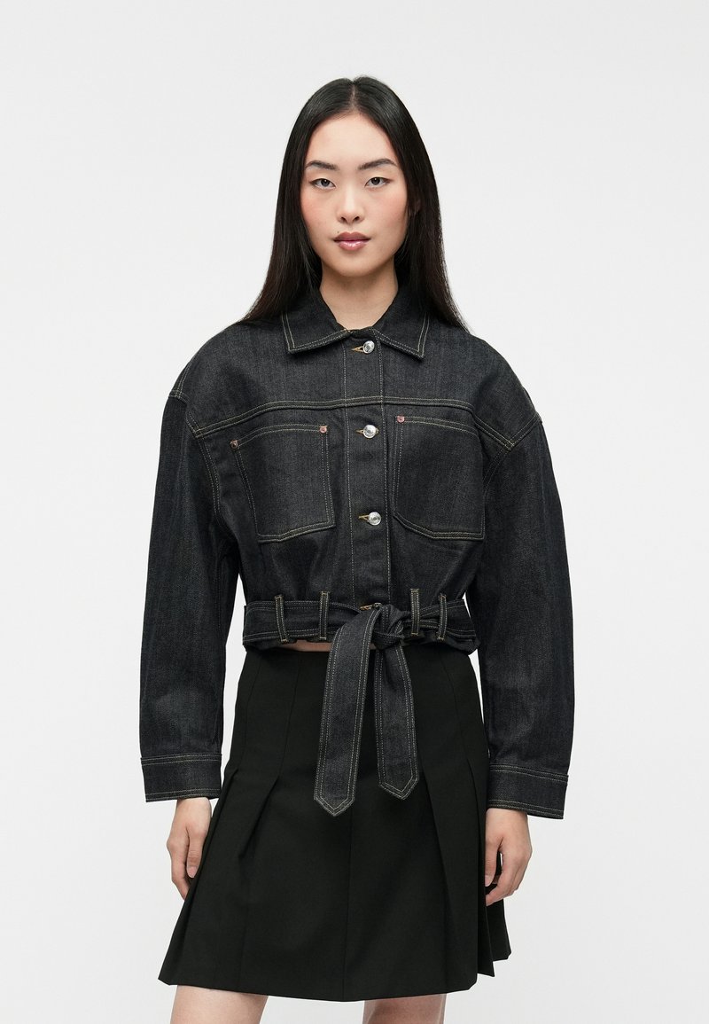 Chaqueta de denim negro con un diseño corto, botones y dos bolsillos frontales; presenta una cintura atada y costuras amarillas en contraste.