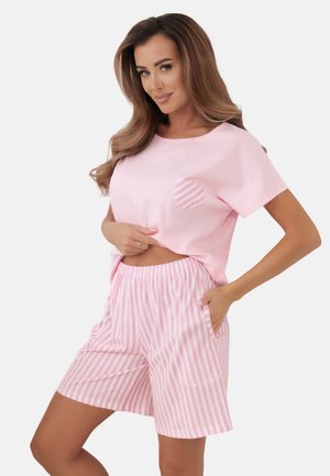 Kobieta ubrana w różowy t-shirt z krótkim rękawem z detalem w postaci paskowanego serca oraz pasujące różowo-białe pionowo paskowane szorty, pozuje z jedną ręką w kieszeni.
