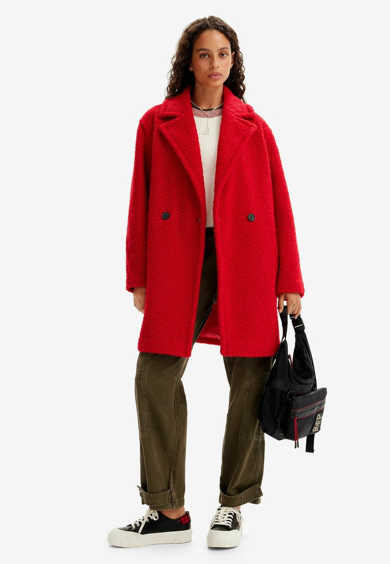 Cappotto rosso peloso doppiopetto con ampi revers e bottoni neri, abbinato a pantaloni verdi oliva e sneakers nere con suole bianche.