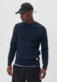 Navy ribbed knit sweater with a crew neckline and long sleeves, paired with a black cap featuring a logo. Black pants complete the outfit.