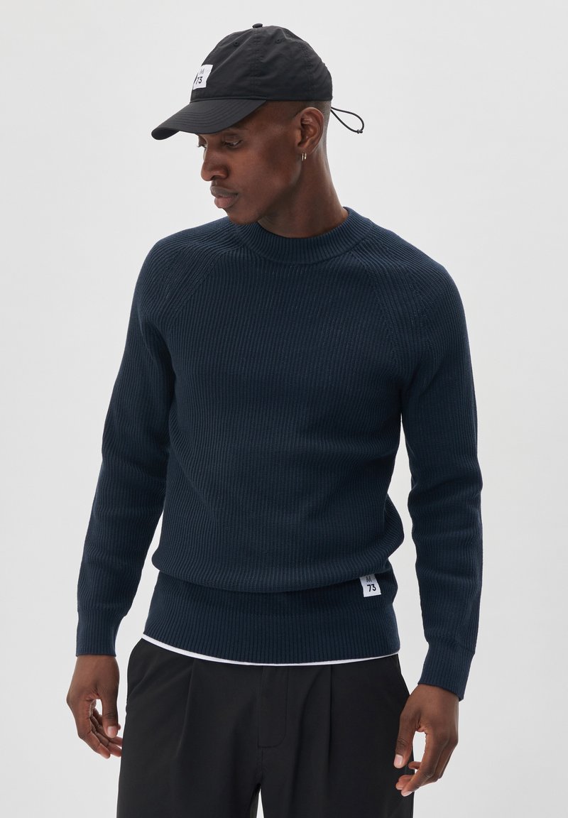 Navy ribbed knit sweater with a crew neckline and long sleeves, paired with a black cap featuring a logo. Black pants complete the outfit.