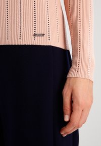 Top a maniche lunghe rosa a coste, con righe orizzontali e piccoli ritagli, dotato di un'etichetta del marchio argentata, abbinato a un tessuto blu scuro.