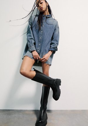 Tânără cu cozi lungi împletite, purtând o cămașă din denim supradimensionată și cizme negre masive până la genunchi, pozând în fața unui perete alb simplu.