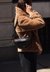 Zwarte leren schoudertas met gouden rits en studaccenten. Draagt samen met een lichtbruine fluffy jas en zwarte broeken. Daglichtinstelling.