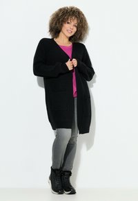 Svart stickad cardigan med texturerad tyg, två fickor fram, parat med en rosa topp, grå jeans och svarta snörkängor.