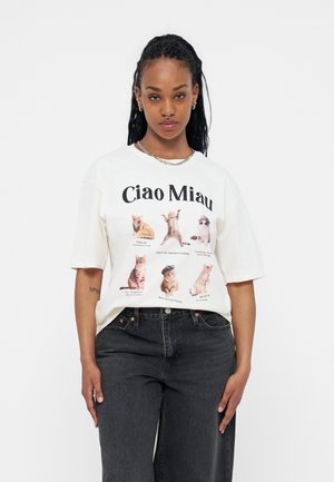 Woman with braided hair wears white T-shirt featuring six playful cats and text "Ciao Miau" paired with black jeans against plain background.
