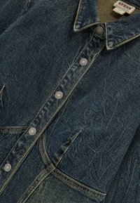 Denimjasje met een donkerblauwe wassing, knoopsluiting aan de voorkant, spitse kraag en metalen knopen. Zichtbare stiksels en zakken verbeteren het ontwerp.