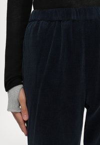 Main reposant à côté d’un pantalon en velours côtelé noir à taille élastique, portant un haut à manches longues noir et une manche intérieure grise avec un trou pour le pouce.