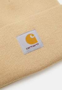 Beežist kootud beanie, millel on sooneline tekstuur ja ruudukujuline patch, millel on kollane logo ja sõna "carhartt" tumedates tähtedes.