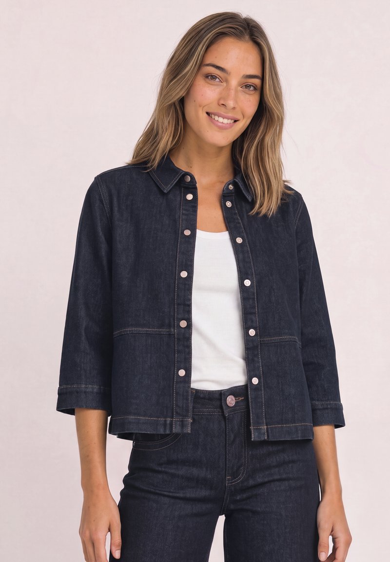 Jeune femme portant une veste en jean bleu foncé et un jean avec un haut blanc, souriante, debout devant un fond clair uni.