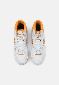 Une paire de baskets New Balance 650 blanches et orange avec des lacets, vue de dessus sur un fond blanc uni.