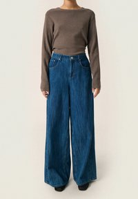 Braunes langärmliges Oberteil, kombiniert mit hoch taillierten, weiten blauen Jeans aus Denim, die zwei frontale Taschen und einen Metallknopfverschluss aufweisen.