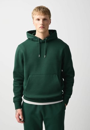 Junger Mann trägt einen dunkelgrünen Hoodie mit Fronttaschen und passenden Hosen und steht vor einem schlichten hellen Hintergrund.