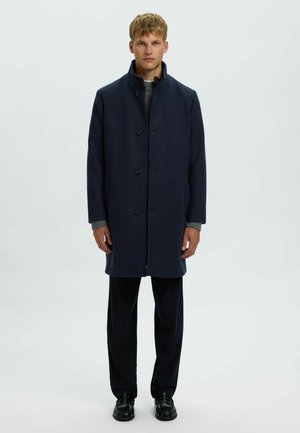 Jeune homme debout face à l'avant, portant un manteau bleu foncé boutonné, un pull gris, un pantalon noir et des chaussures noires sur un fond uni.
