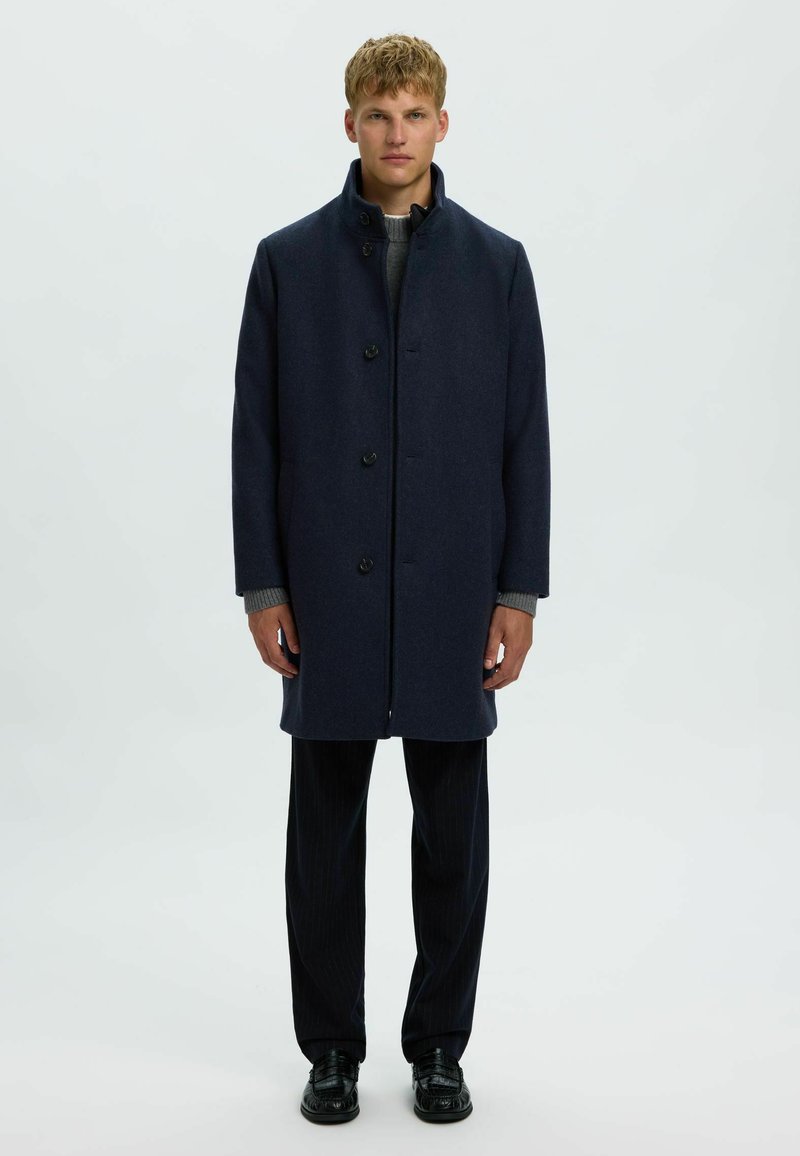 Jeune homme debout face à l'avant, portant un manteau bleu foncé boutonné, un pull gris, un pantalon noir et des chaussures noires sur un fond uni.