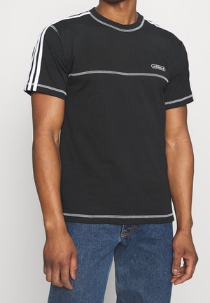 Man draagt een zwart Adidas T-shirt met witte contrastnaden en strepen op de mouwen, gecombineerd met een blauwe spijkerbroek, staand tegen een effen achtergrond.