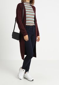 Długi bordowy kardigan na paskowym dzianinowym topie, w połączeniu z ciemnymi dżinsami i białymi trampkami. Dopełnieniem stroju jest mała czarna torba na ramię.