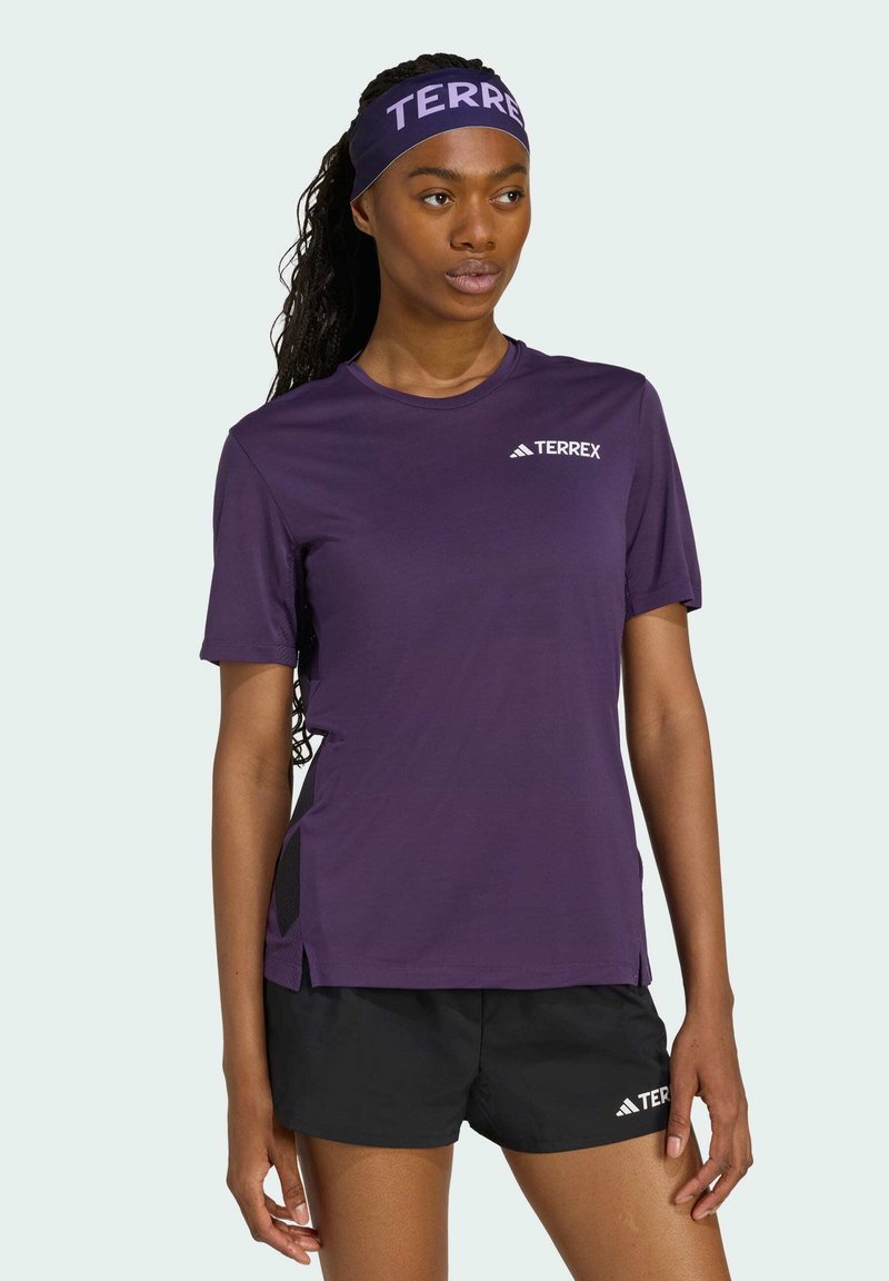 Camiseta deportiva morada de manga corta, con paneles de malla negra a los lados y el logo blanco "TERREX". Combinada con pantalones cortos negros.