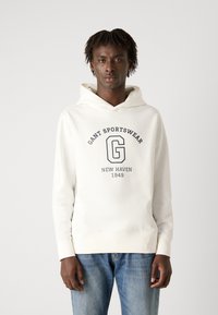 Sudadera blanca con un gráfico frontal que dice "GANT SPORTSWEAR NEW HAVEN 1949". Cuenta con capucha ajustable y puños acanalados, combinada con jeans azules.