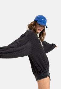 Black textured sweatshirt with oversized sleeves and a white drawstring. Worn with dark denim shorts and a blue baseball cap.
