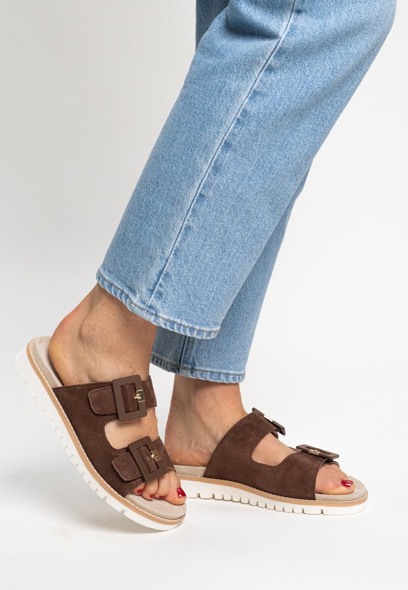 Sandales glissées en daim marron avec double boucle, semelle texturée blanche, design à bout ouvert et une semelle beige, associées à un jean bleu court.