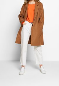 Frau trägt einen braunen Mantel über einem orangen Oberteil, weiße gekürzte Jeans und weiße Turnschuhe, steht mit einer Hand in der Tasche.