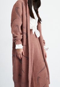 Översized rosa fleecekofta med brunt abstrakt mönster, med långa ärmar och öppet fram, parat med en vit knuten skjorta och rosa kjol.