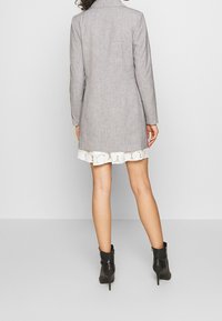 Szary, teksturowany długi żakiet o dopasowanym kroju, w połączeniu z białą sukienką wykończoną koronką i noszony z czarnymi botkami do kostki.