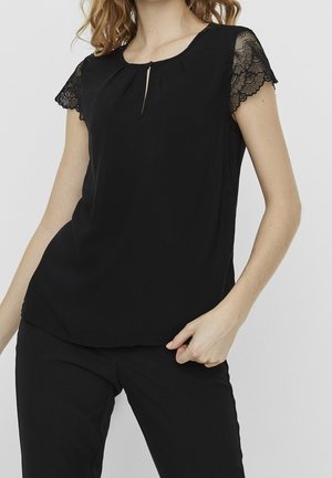 Femme portant un chemisier noir à manches courtes avec des détails en dentelle sur les manches et une petite ouverture devant, associée à un pantalon noir.