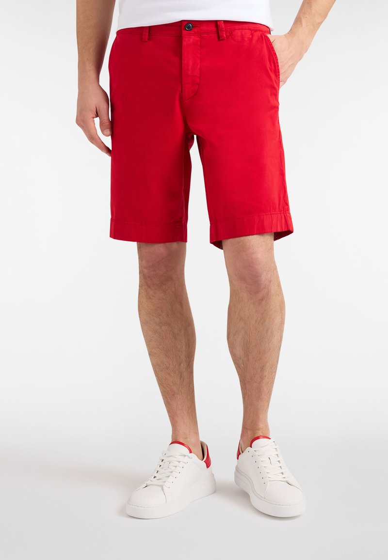 Červené bavlnené šortky so zahnutými lemami, bočnými vreckami a predným zapínaním na gombíky. Sú doplnené bielymi teniskami s červenými akcentmi.