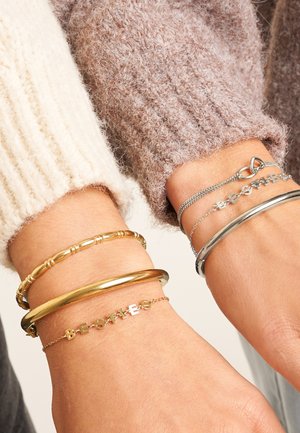 Zwei Handgelenke mit Armbändern, darunter goldene und silberne Armreifen sowie zarte Ketten, die das Wort "BESTIES" bilden, getragen über gemütlichen Strickpullovern.