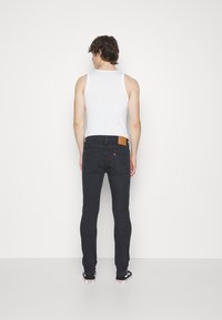 Jeans oscuros de corte ajustado con una etiqueta de cuero en la cintura. El modelo lleva una camiseta sin mangas blanca y zapatillas negras.