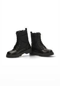 Botas negras hasta el tobillo que presentan un exterior de cuero liso y brillante, un eje acolchado, costuras decorativas y cordones, todo con una suela de goma gruesa.
